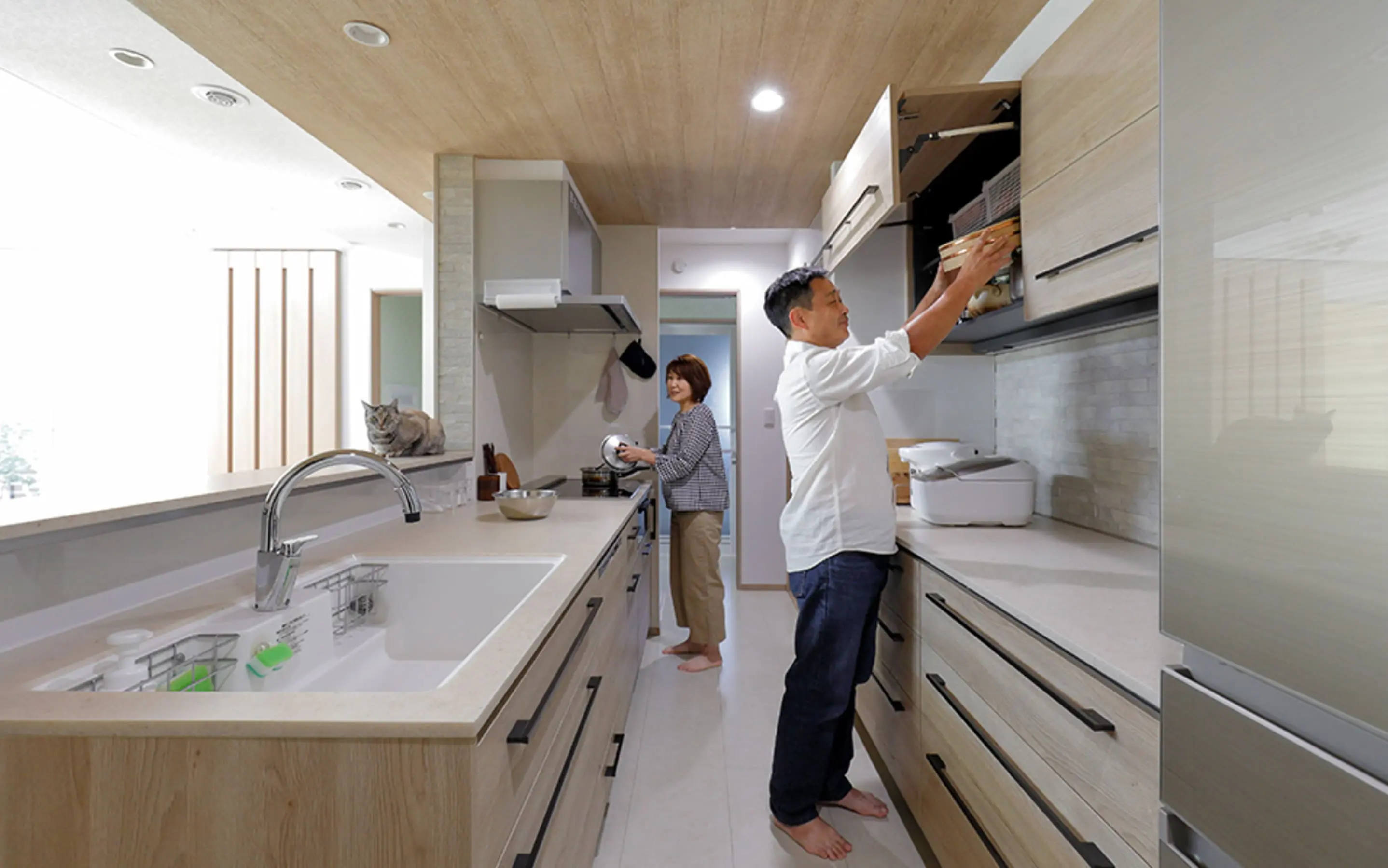 木目調のキッチンで夫婦が料理の準備をしており、猫がカウンター上に座っている温かな日常の一コマ