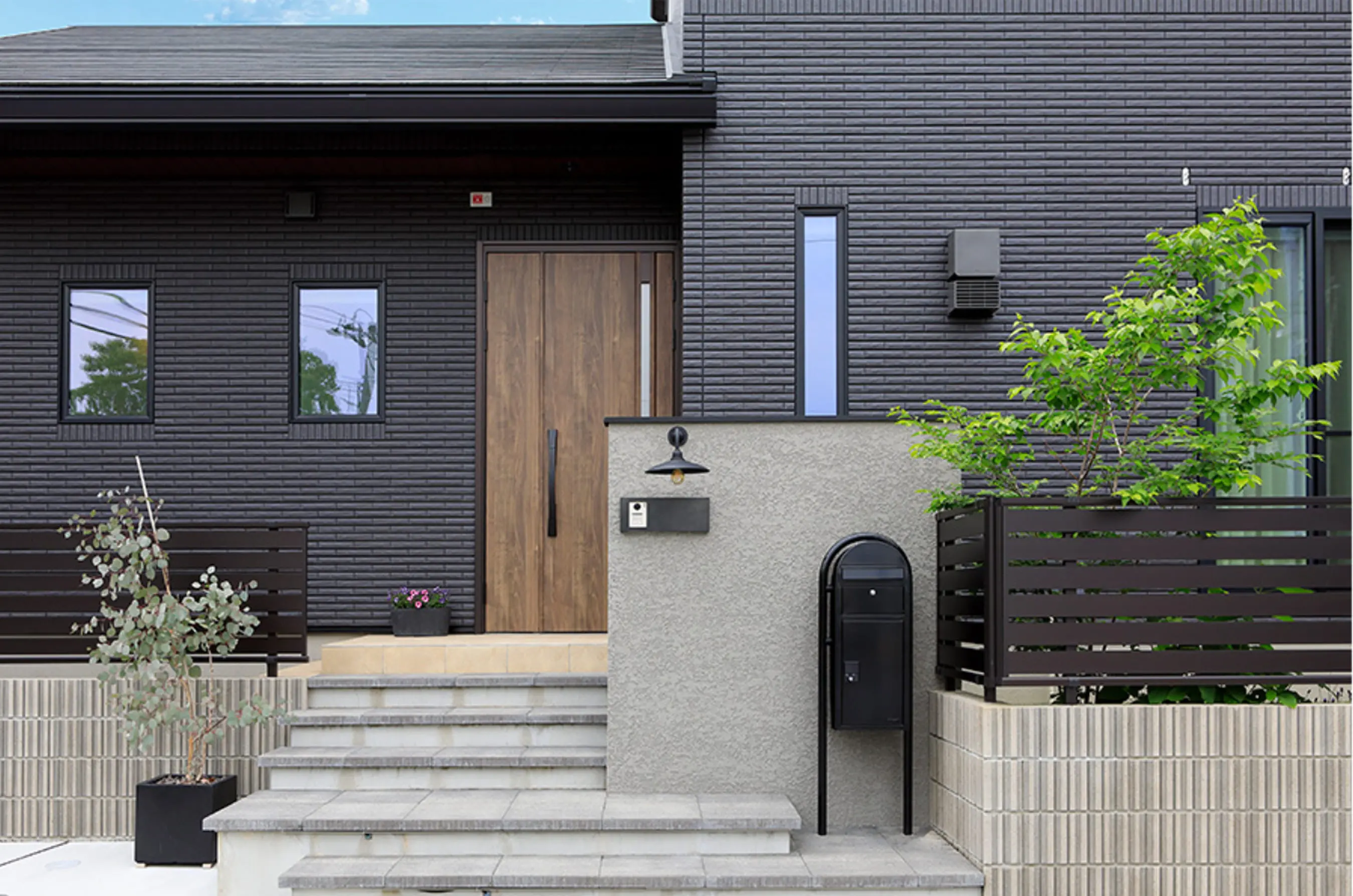 木製ドアと植栽が印象的な玄関まわりで、グレーの塀と黒いポストが調和している