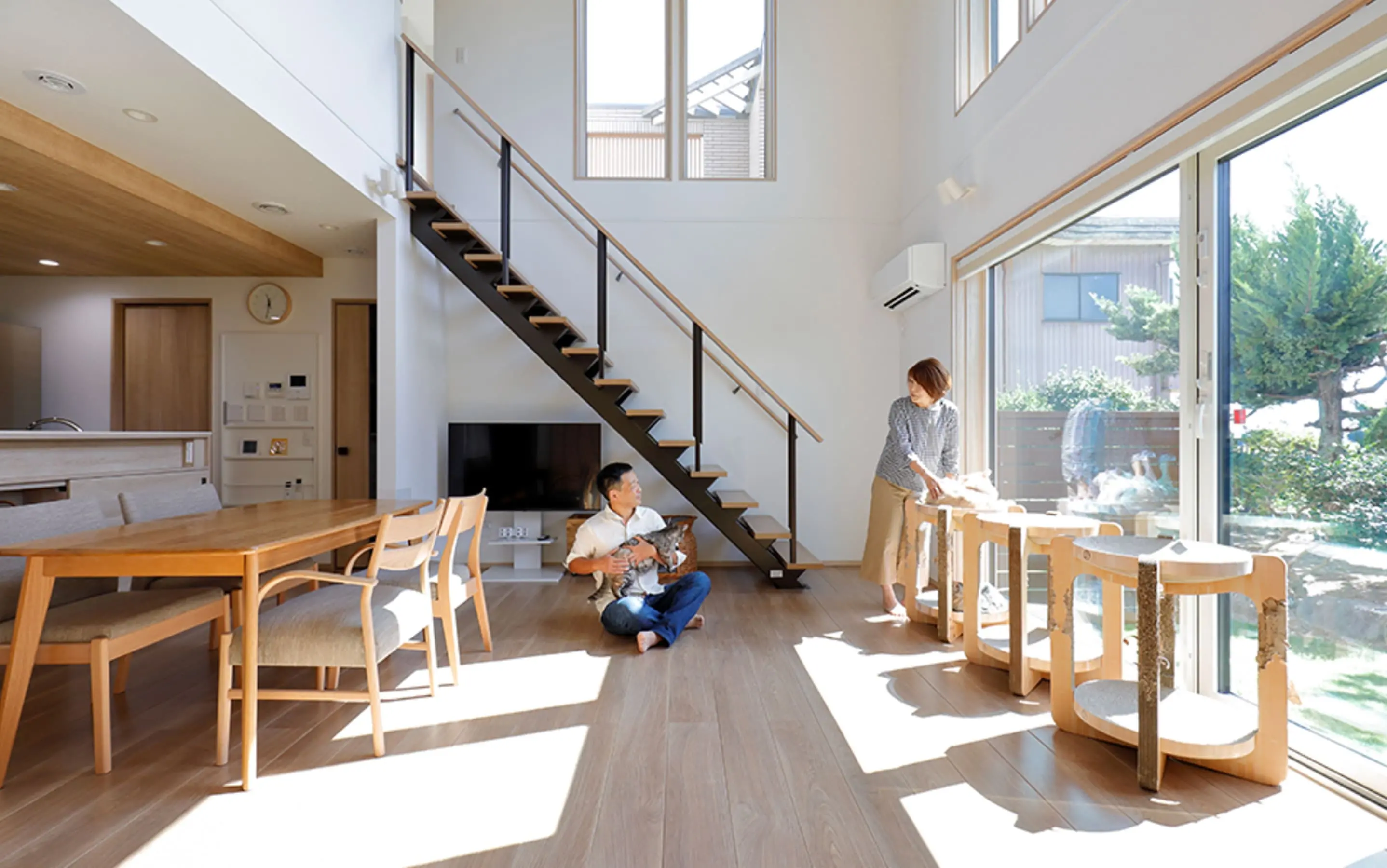 吹き抜けのリビングに大きな窓があり、自然光が差し込む中で夫婦が猫と過ごしている様子