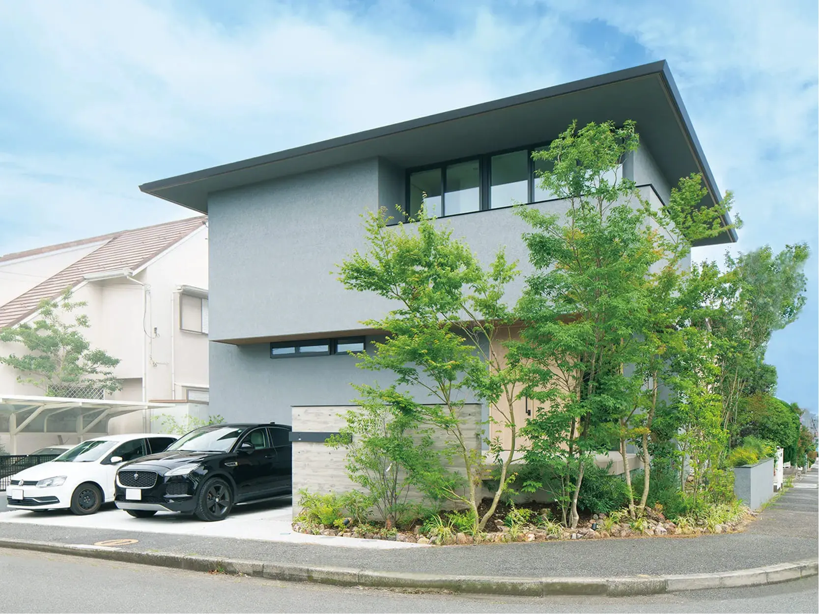 グレーの外壁と深い軒が特徴の二階建て住宅で、植栽と2台の車が並ぶスタイリッシュな外観